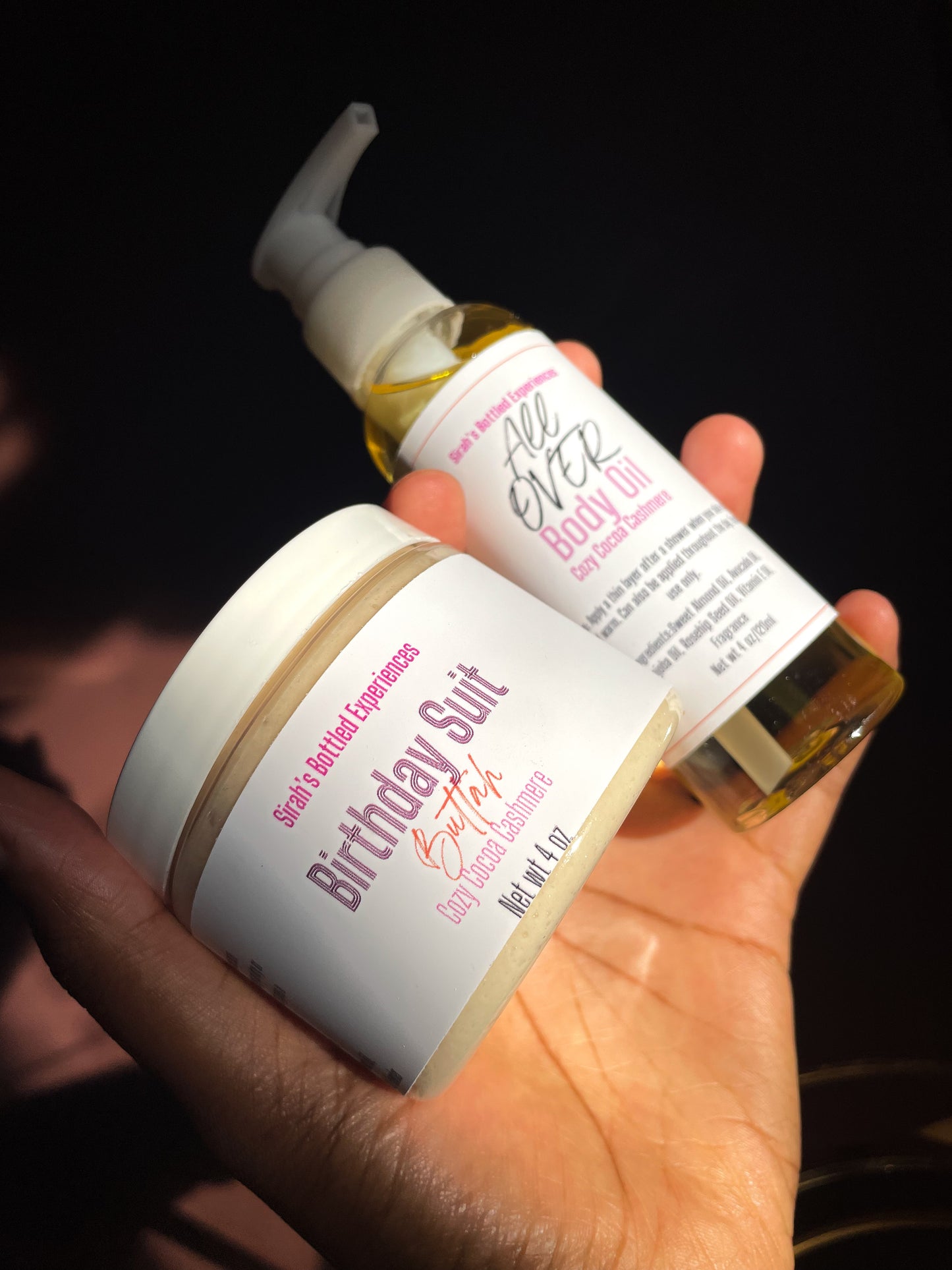 Hand holding a jar and bottle of 'All Over' body oil and a jar of birthday suit butter whipped