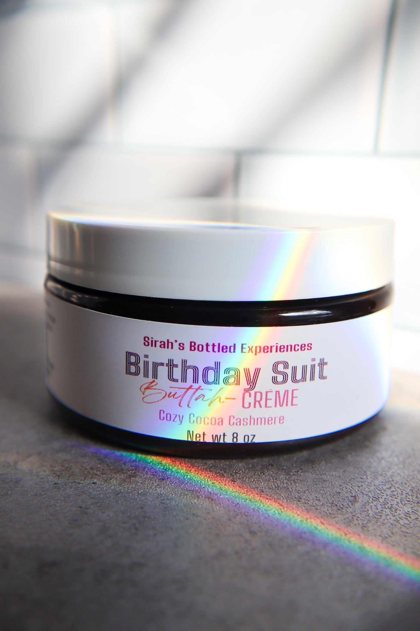 Jar of 'Birthday Suit Butter Creme' with a rainbow effect on a gray surface