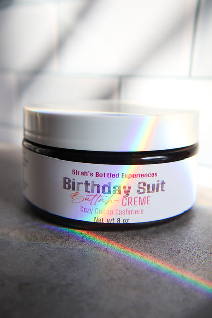Jar of 'Birthday Suit Butter Creme' with a rainbow effect on a gray surface