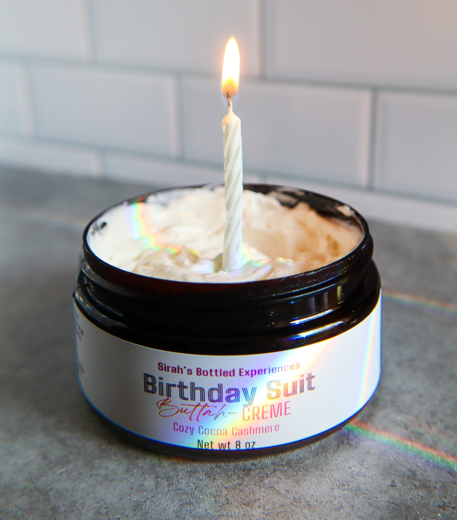 Jar of 'Birthday Suit Butter Creme' with a lit candle inside, on a tiled surface.